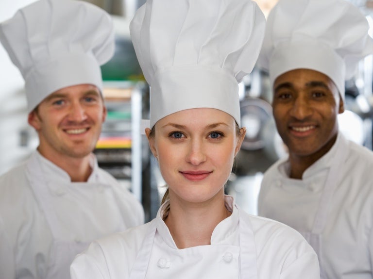In a well-lit kitchen, three cheerful chefs pose together, exuding a sense of teamwork and culinary passion.