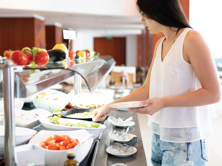 Woman choosing salad at a restaurant buffet counter with bowls of fresh vegetables.