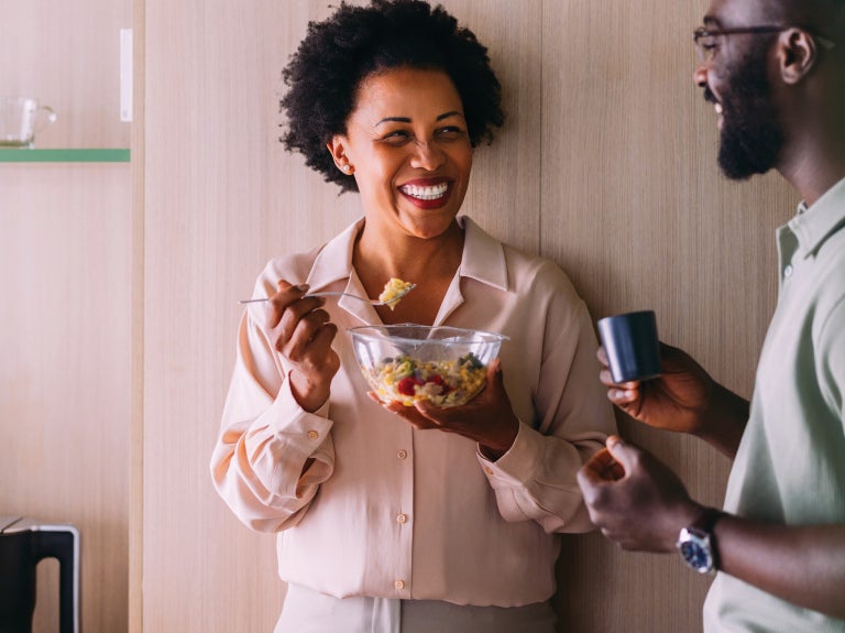 Smiling woman eats salad from a glass bowl while chatting with a man holding a cup in a workplace kitchen.