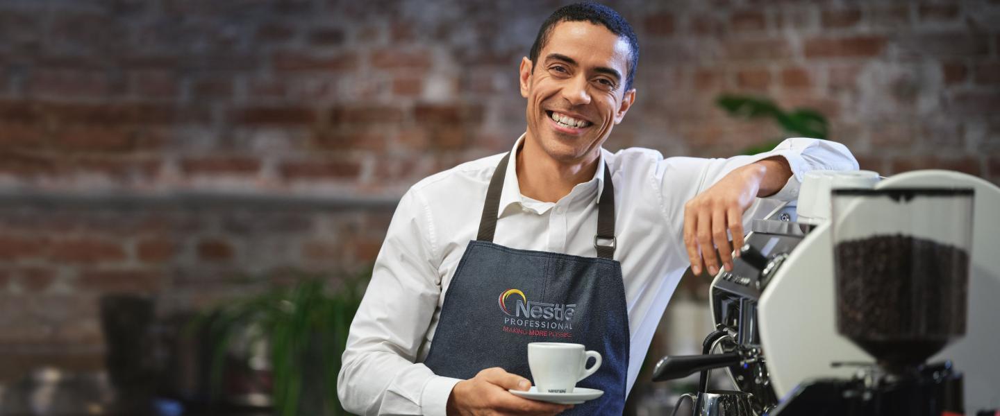 A smiling man in an apron holds a coffee cup, radiating warmth and friendliness.
