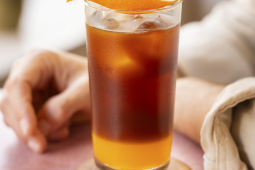A person holding a tall glass of orange iced tea with ice cubes and a slice of orange on the rim.