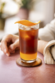 A person holding a tall glass of orange iced tea with ice cubes and a slice of orange on the rim.