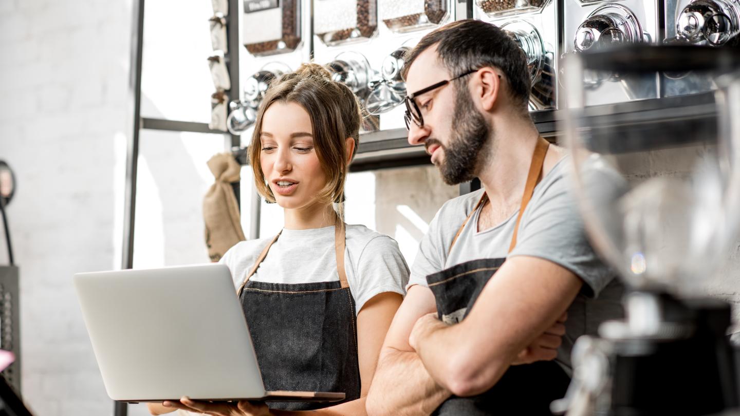 A man and woman in aprons together, looking at a laptop on a table, engaged in a cooking activity.