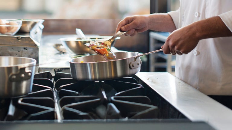 Chef sauteing vegetables in a stainless pan over a gas stove in a commercial kitchen.